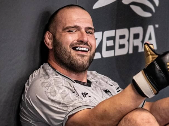 UFC heavyweight Martin Buday sits against the cage with a mouthguard and gloves, smiling after a fight. The text explains factors behind martin buday release, including contract expiry, fighting style, roster management, and marketability.
