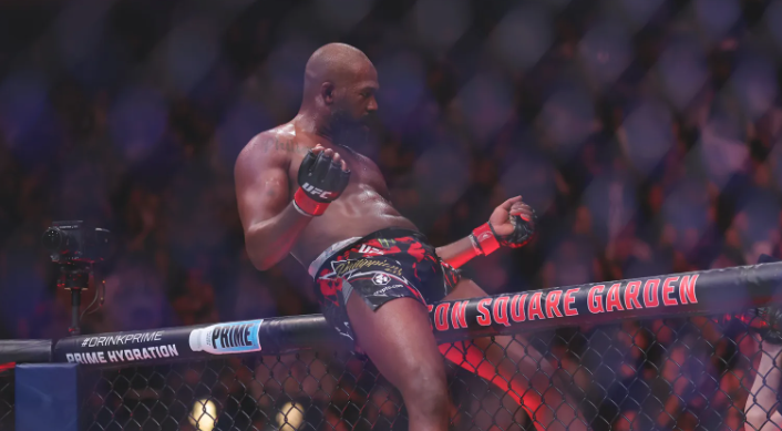 Jon Jones stands atop the UFC cage at Madison Square Garden, flexing his fists in celebration after a victory, embodying both triumph and the weight of his controversial legacy.
