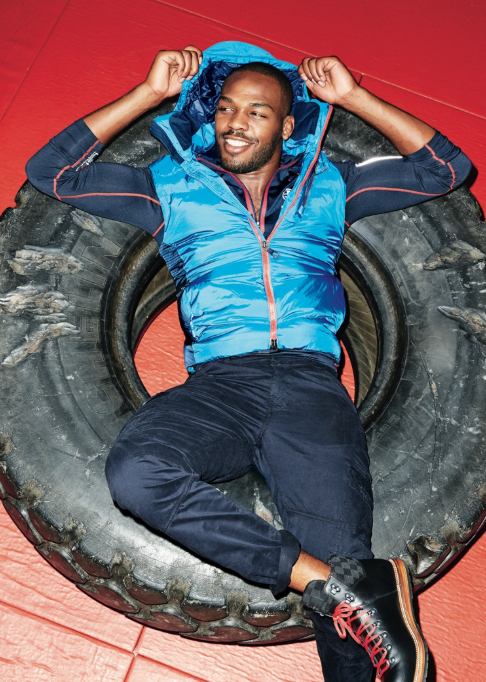 Jon Jones smiles while resting inside a large training tire, wearing a blue athletic vest and boots, reflecting a relaxed moment during his post-retirement phase.
