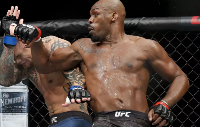 Jon Jones throws a powerful punch during a UFC fight, wearing black shorts with the UFC logo. The text highlights his June retirement as undisputed light heavyweight champion and hints at a possible 2026 comeback.
