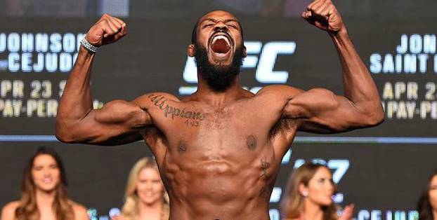 Jon Jones flexes and roars on stage at UFC weigh-ins, showing his muscular physique and tattoos. The text discusses his possible return to competition and Dana White’s doubts about a future fight.
