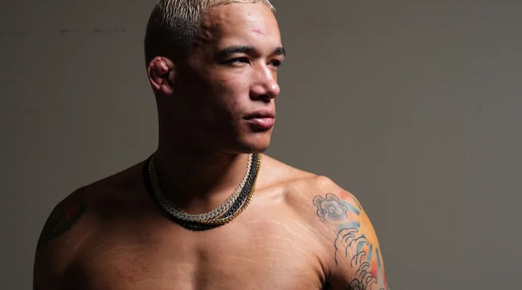 Bryan Battle poses shirtless against a neutral background, looking off to the side with a serious expression, wearing a chain necklace and showing tattoos on his shoulder and chest.